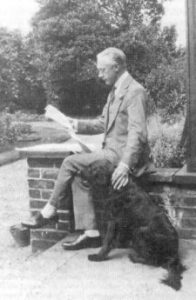 F.M. Alexander sitting outside reading the paper with hand on dog at his side